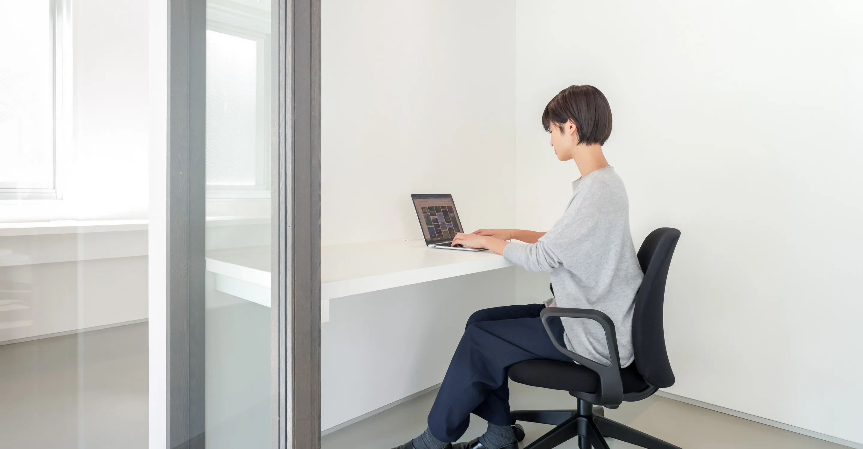 写真。白い壁とデスクの前にオフィスチェアに座ってパソコンを打ち込んでいる女性