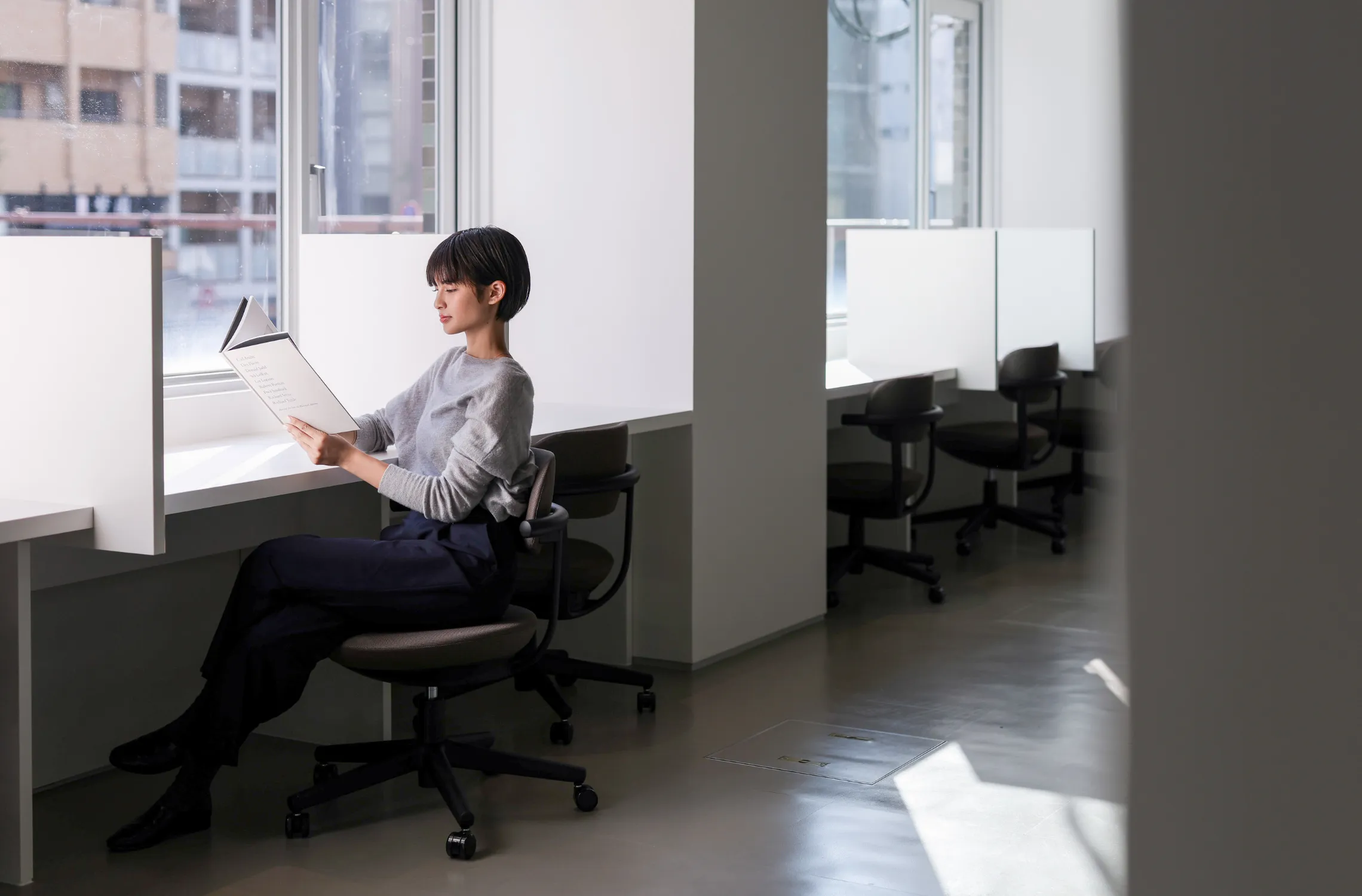 写真。窓際の仕切りのあるデスクで書類を手に読んでいる女性